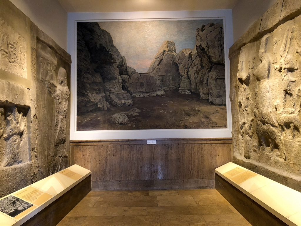 A museum exhibit featuring a large landscape painting of rocky terrain between two walls adorned with ancient stone carvings. The display includes informational plaques on the benches below the carvings.