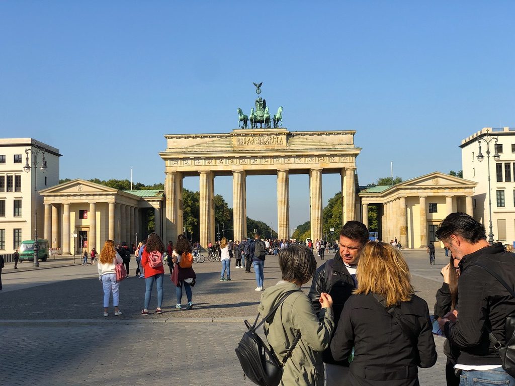 People gather near the iconic Brandenburg Gate in Berlin on a sunny day. The neoclassical monument has a Quadriga statue on top, with clear blue skies overhead. Visitors are walking and taking photos, framed by historic buildings on either side.