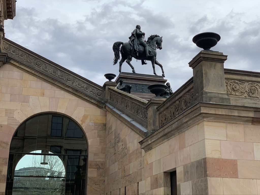 A historic stone building with architectural details and an arched glass window. A statue of a man on horseback is prominently displayed on the rooftop. The sky is overcast with clouds.