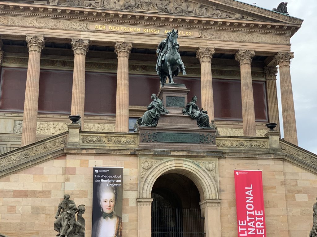 The image shows the entrance of the Alte Nationalgalerie in Berlin. A grand staircase leads up to the building, which has tall columns and a large statue of a rider on horseback prominently displayed in front. Banners adorn the facade.