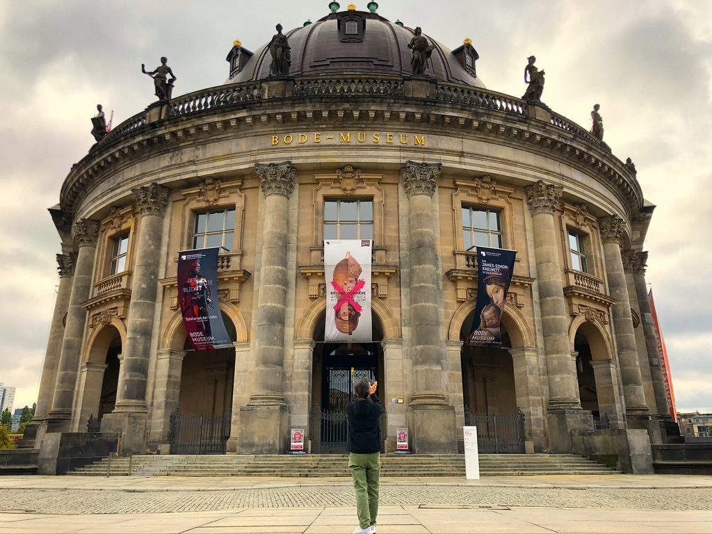 A person stands in front of the Bode Museum in Berlin, capturing a photo. The building features a grand dome, statues on top, and large banners on the facade. The sky is overcast, adding dramatic contrast to the scene.