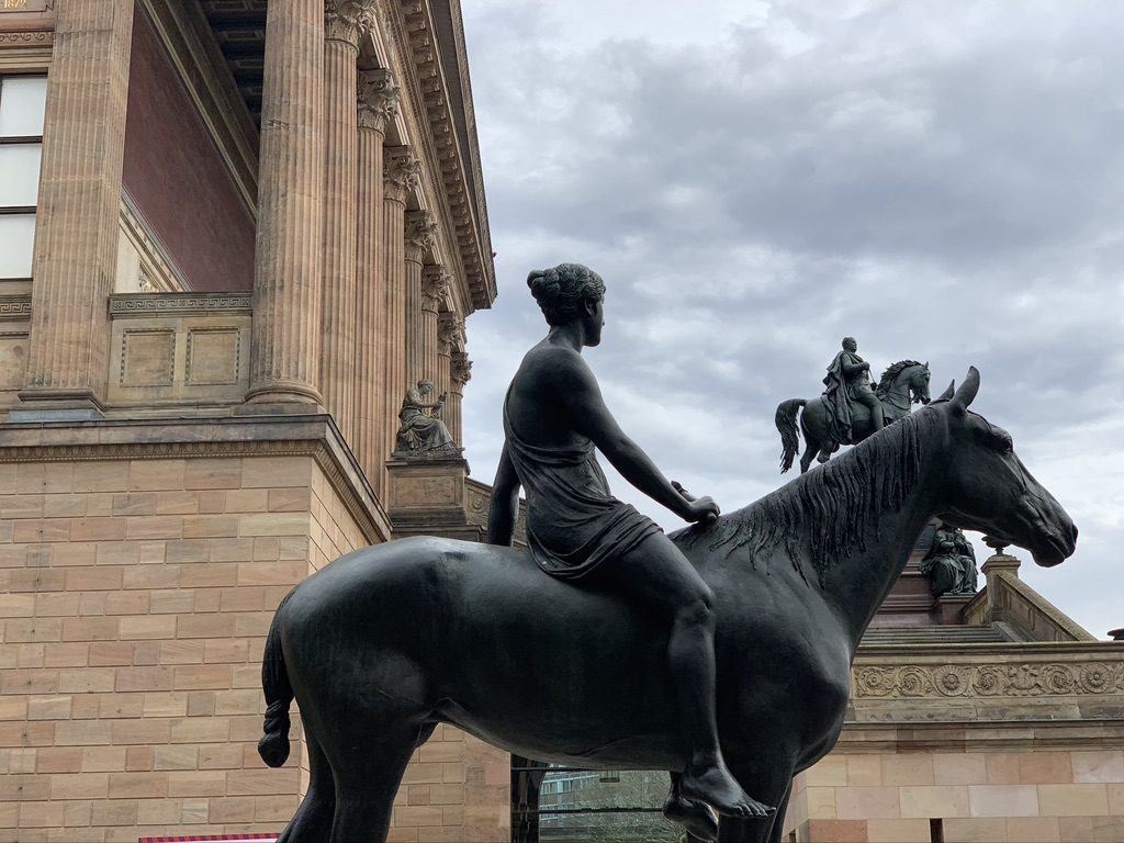 A statue of a rider on horseback is seen in front of a neoclassical building with tall columns. Another equestrian statue is visible in the background. The sky is overcast.