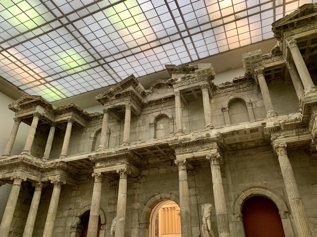 A large ancient stone structure with ornate columns and archways under a modern glass ceiling. The architecture features detailed carvings and varying depths, with some openings leading to further rooms.
