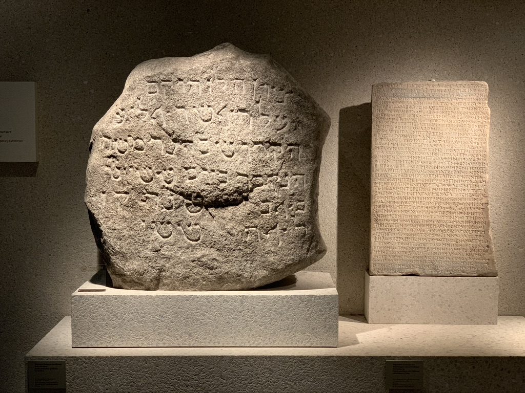 A display of two ancient stone tablets with engraved inscriptions, one irregularly shaped with bold script, the other rectangular with fine writing, set against a dimly lit museum background.