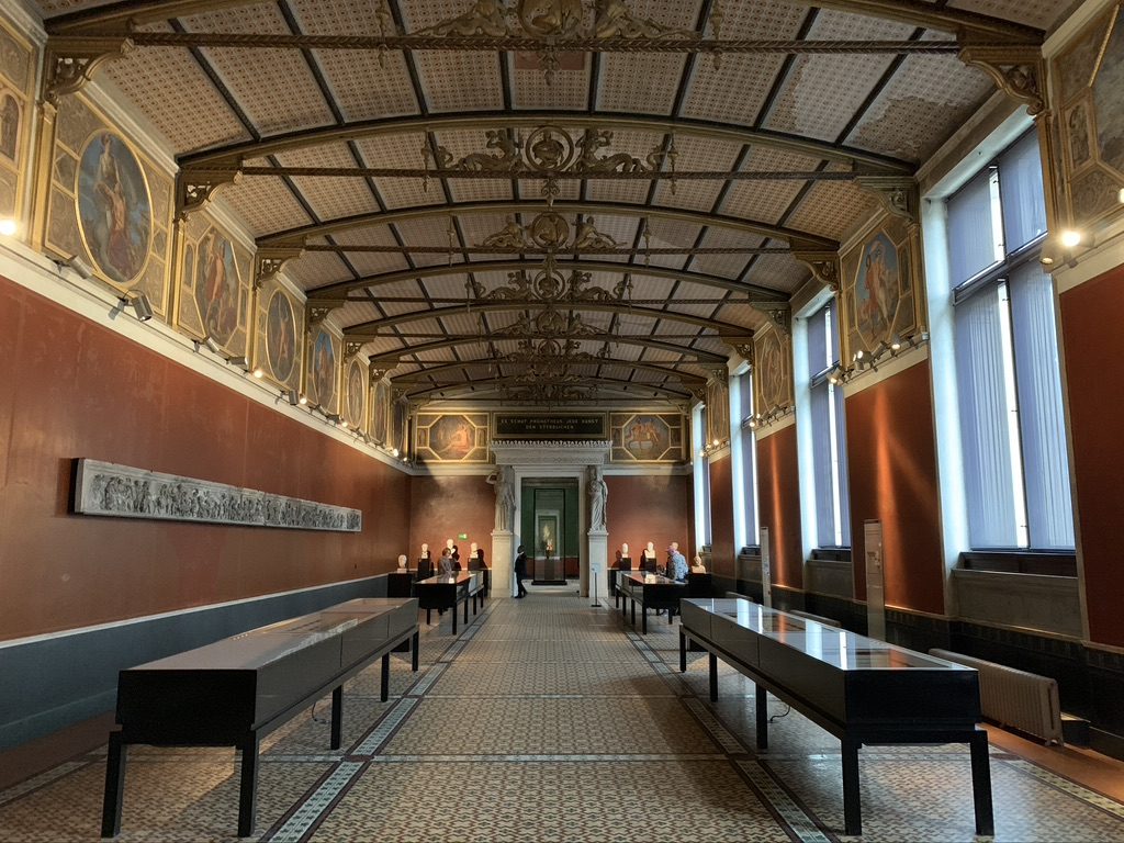An ornate, high-ceilinged room with red walls and decorative paintings along the upper section. It features long, black display cases arranged in two rows and a statue at the far end. Natural light streams in through tall windows on the right.