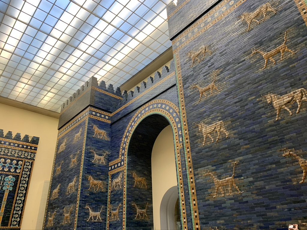 The image shows the Ishtar Gate, a large ancient structure adorned with blue glazed bricks and decorated with images of bulls and dragons. It features an arched entryway and is lit by a skylight above.