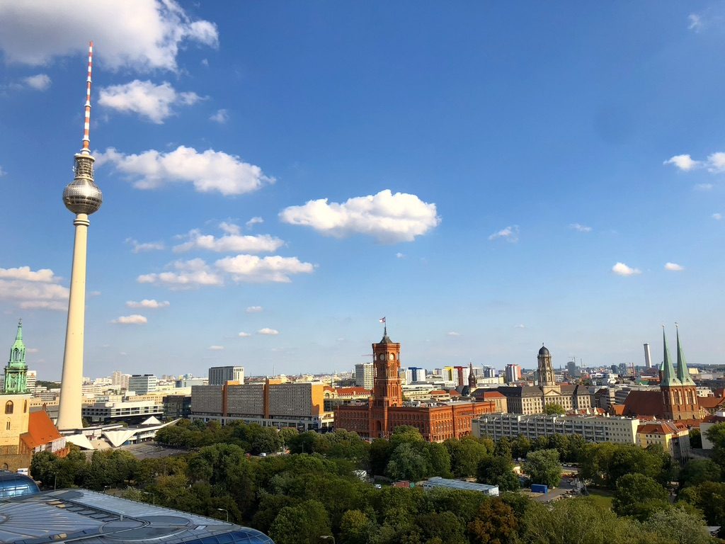 A panoramic view of Berlins cityscape with the iconic Fernsehturm tower on the left, the historic red-brick Rotes Rathaus in the center, and various buildings and greenery under a blue sky scattered with clouds.
