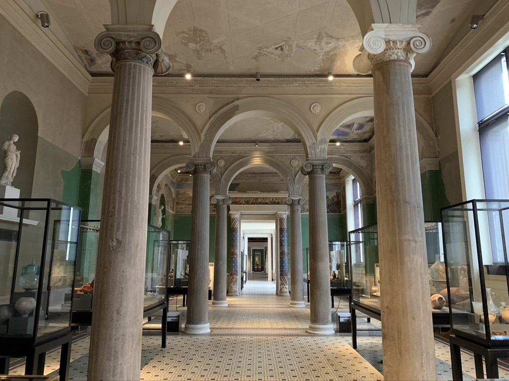A grand museum corridor with tall marble columns and arched ceilings leading to a distant doorway. Display cases filled with artifacts line the walls, and statues are visible in niches. The floor is patterned, enhancing the classical ambiance.