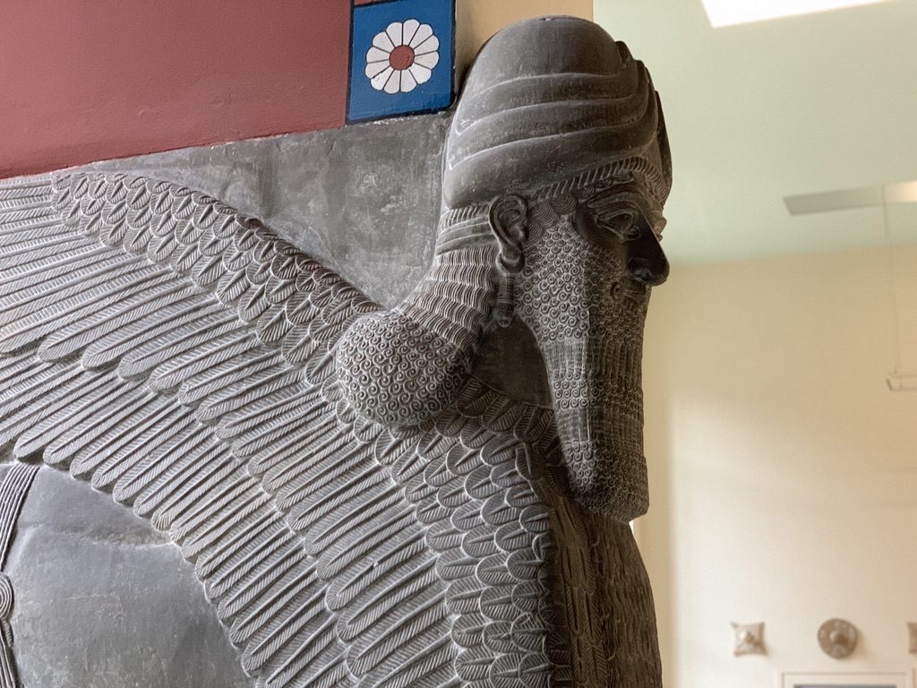 Ancient Assyrian stone relief depicting a winged, bearded figure wearing a horned headdress. The carving shows intricate details, such as feathers and curls, with a colored decorative border featuring a flower above.