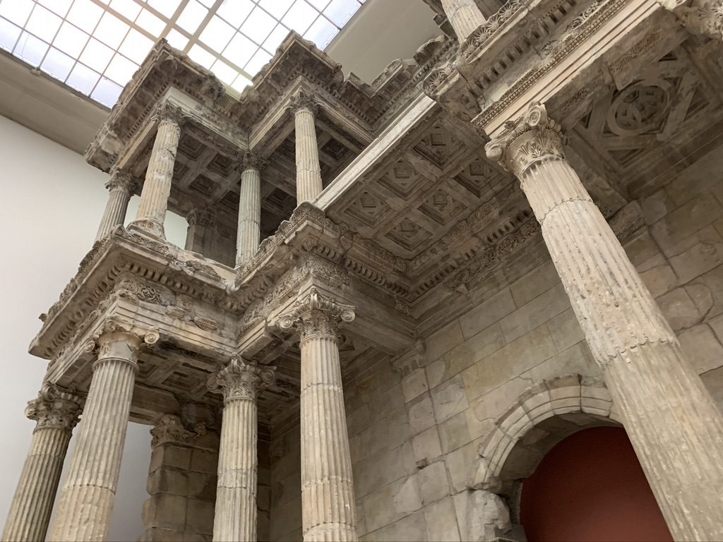 The image shows a detailed view of an ancient Roman architectural structure with tall, ornate columns, intricate carvings, and an arched doorway. The ceiling features a geometric pattern, and natural light streams through a skylight above.