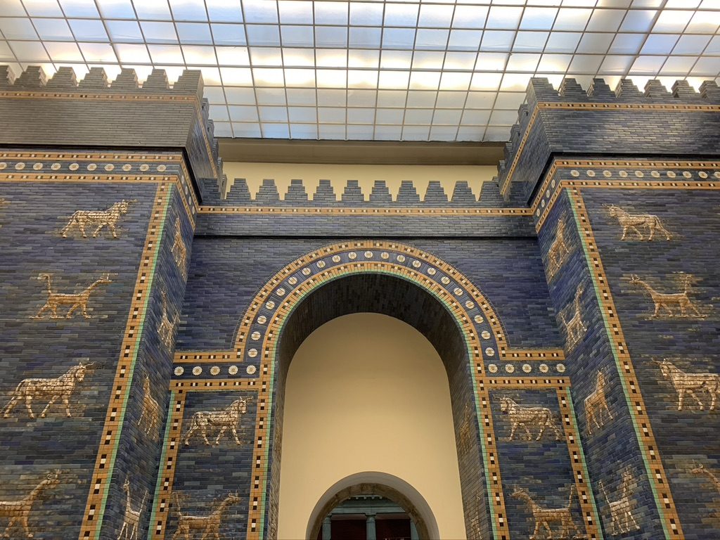 The image shows a section of the reconstructed Ishtar Gate, featuring blue glazed bricks with decorative rows of animal reliefs, including dragons and bulls. The scene is under a glass-paneled ceiling.