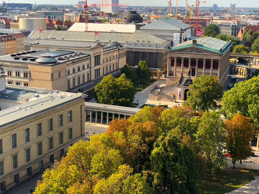 Aerial view of a tree-lined area in a city featuring several classical buildings, one with columns. The scene includes green spaces, a bridge-like structure, and cranes visible in the background under a clear sky.