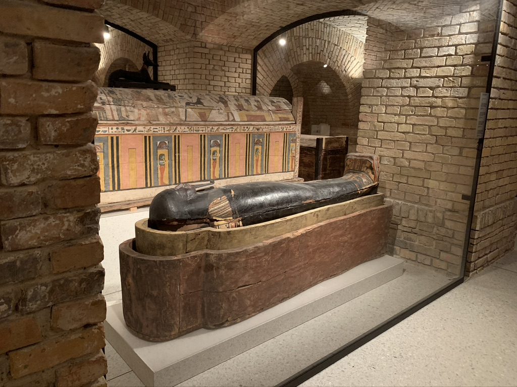 An ancient Egyptian sarcophagus displayed in a dimly lit museum setting, placed within a brick-walled room. The outer lid is painted and intricately decorated, while the inner casket is exposed, revealing a dark, solid structure.