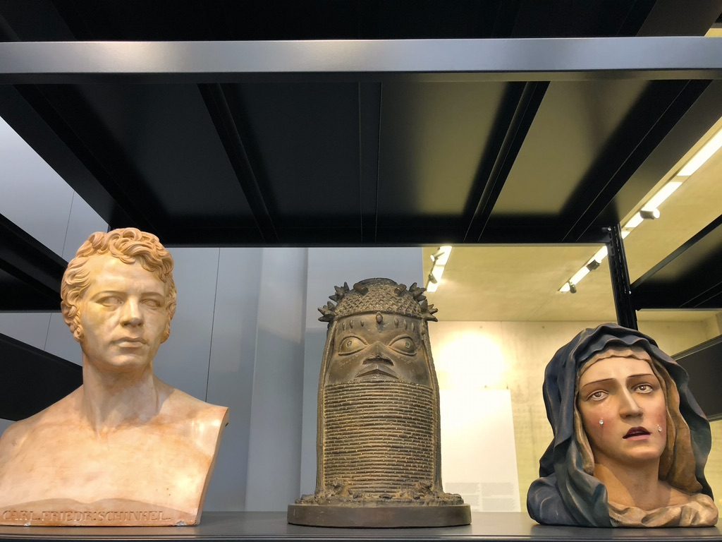 Three sculptures displayed on a shelf: a marble bust of a mans head, a bronze Benin plaque featuring a head with intricate detailing, and a painted bust of a woman wearing a hooded garment, appearing somber.