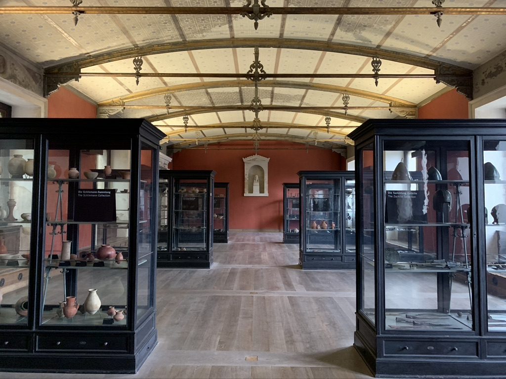 A museum room with several glass display cases filled with various artifacts, including pottery and tools. The room has a high, ornate ceiling and red walls, with wooden floors and lighting highlighting the displays.