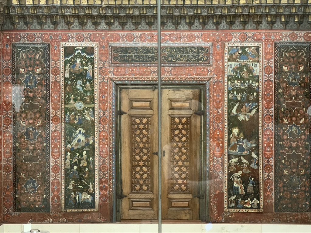 Ornate wooden doors set in an intricate red and gold decorative wall, featuring detailed paintings and geometric patterns. The artwork includes various figures and scenes, surrounded by elaborate floral motifs.