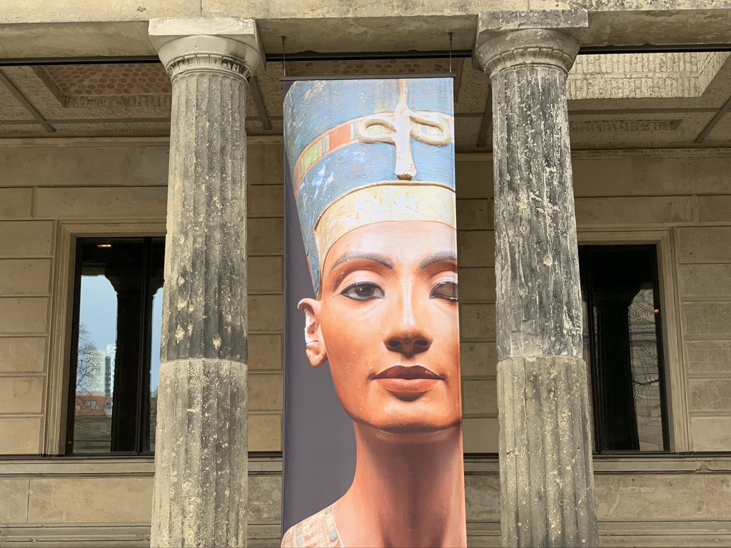 A large vertical banner depicting an ancient Egyptian bust, possibly Nefertiti, is displayed between two weathered stone columns of a historical building. The banner shows a colorful, detailed image of the bust against a gray background.
