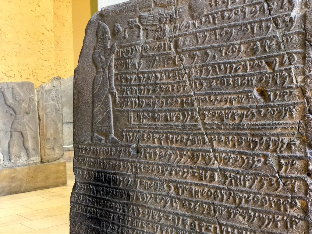 Ancient stone slab with engraved text and figures. The left side depicts a standing figure, while the rest is covered in uniform script. A partially visible relief is in the background. Stone walls form the setting.