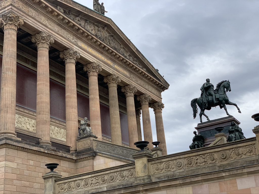 A neoclassical building with tall columns stands under a cloudy sky. A statue of a person on horseback is prominently displayed in front of the building on a raised pedestal. Stone statues are positioned around the structure.