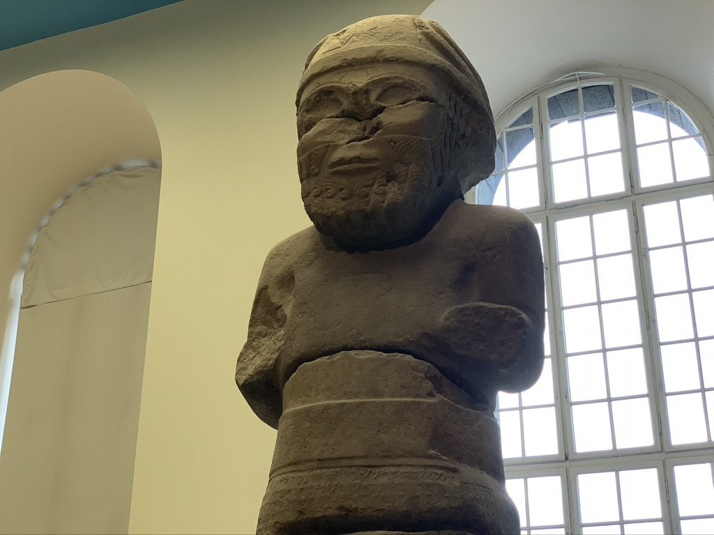 Ancient stone statue with a bearded figure wearing a hat or helmet, standing in front of large, arched windows that allow natural light to illuminate the sculpture. The setting suggests an indoor museum or historical exhibit.
