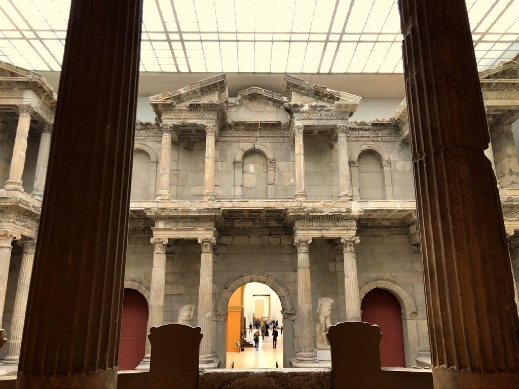 Ancient stone building with columns and arches, viewed from inside. The structure has multiple levels and detailed carvings. Natural light filters through a glass ceiling above. People are visible in the background through an archway.